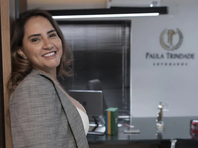 Dra. Paula Trindade sorrindo no escritório, com o logotipo Paula Trindade na parede ao fundo.
